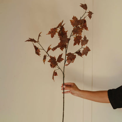 A single artificial-looking branch with brown leaves held by a person's hand against a plain off-white wall.