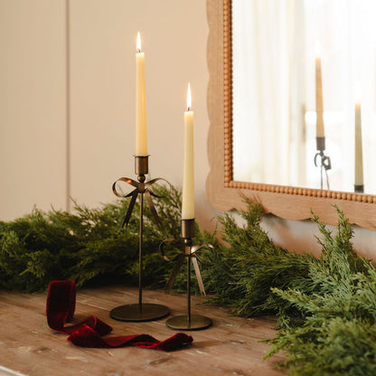 Two candlesticks with lit candles on a wooden surface with greenery and a mirror in the background.