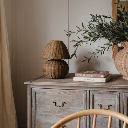 Woven rattan table lamp styled on a rustic wooden sideboard with olive branches, neutral decor, and warm natural tones in a cozy modern interior