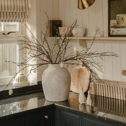 A realistic artificial pussy willow stem in a white vase on a kitchen countertop, surrounded by various home decor items.