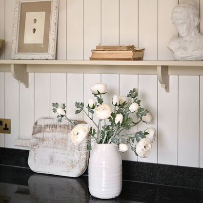 White vase with flowers on a shelf against a white paneled wall.