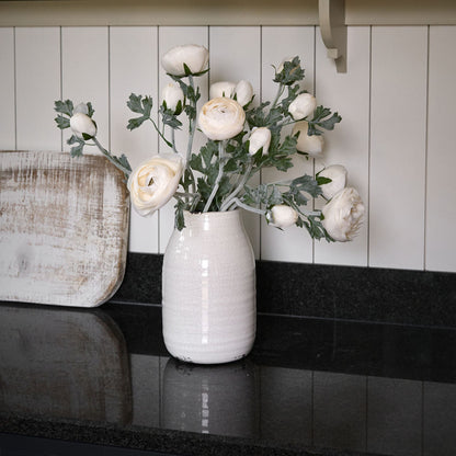 A white ceramic vase with a natural, organic shape, placed on a kitchen counter. The vase contains soft white ranunculus flowers, and is positioned next to a statue and a shelf with books.