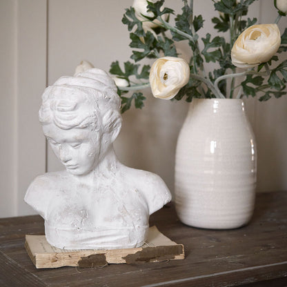 A stone effect female bust icon on a wooden surface, with a cream color and a floral arrangement in the background.