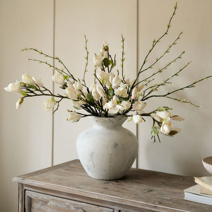 A white, antique-style vase with a Greek design, displayed on a wooden sideboard filled with artificial floral arrangement.