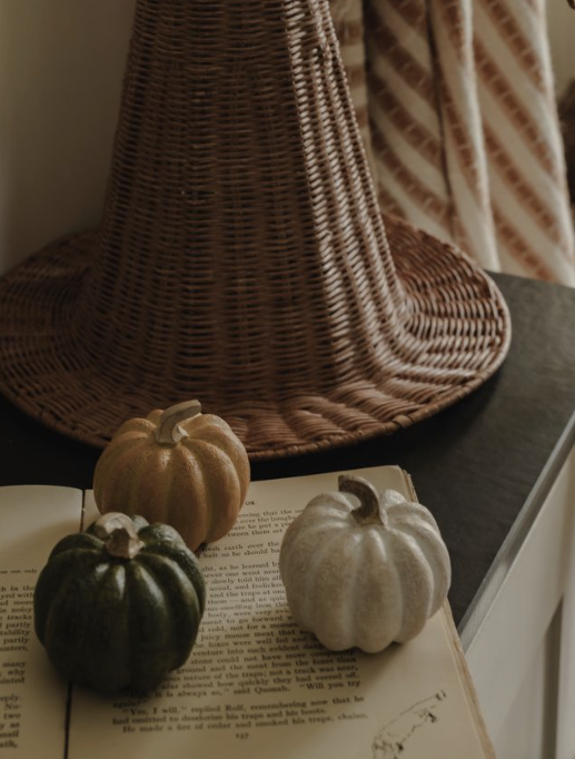 Three decorative miniature pumpkins in dark green, muted gold, and textured off-white are arranged on an open book, with a large woven witch hat in the background.
